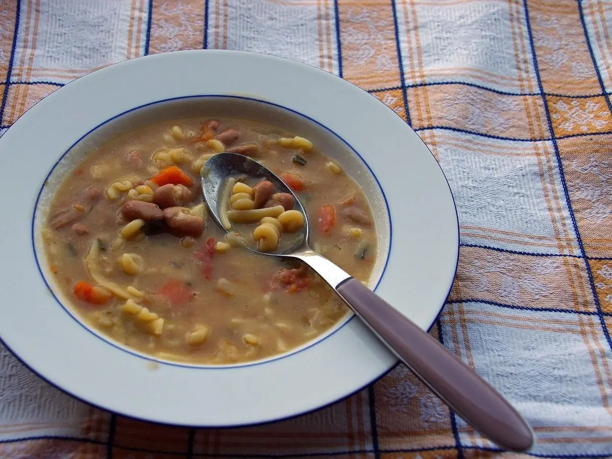 Plato hondo de sopa de pasta con alubias, tomate y verduras con cuchara sobre mantel de cuadros