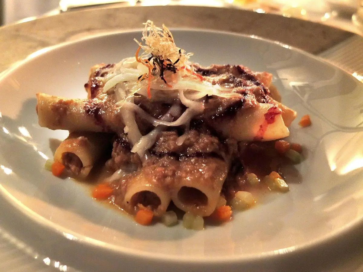 Pasta tubular con ragú de carne y cebolla, cubierta con queso rallado en un plato hondo gris
