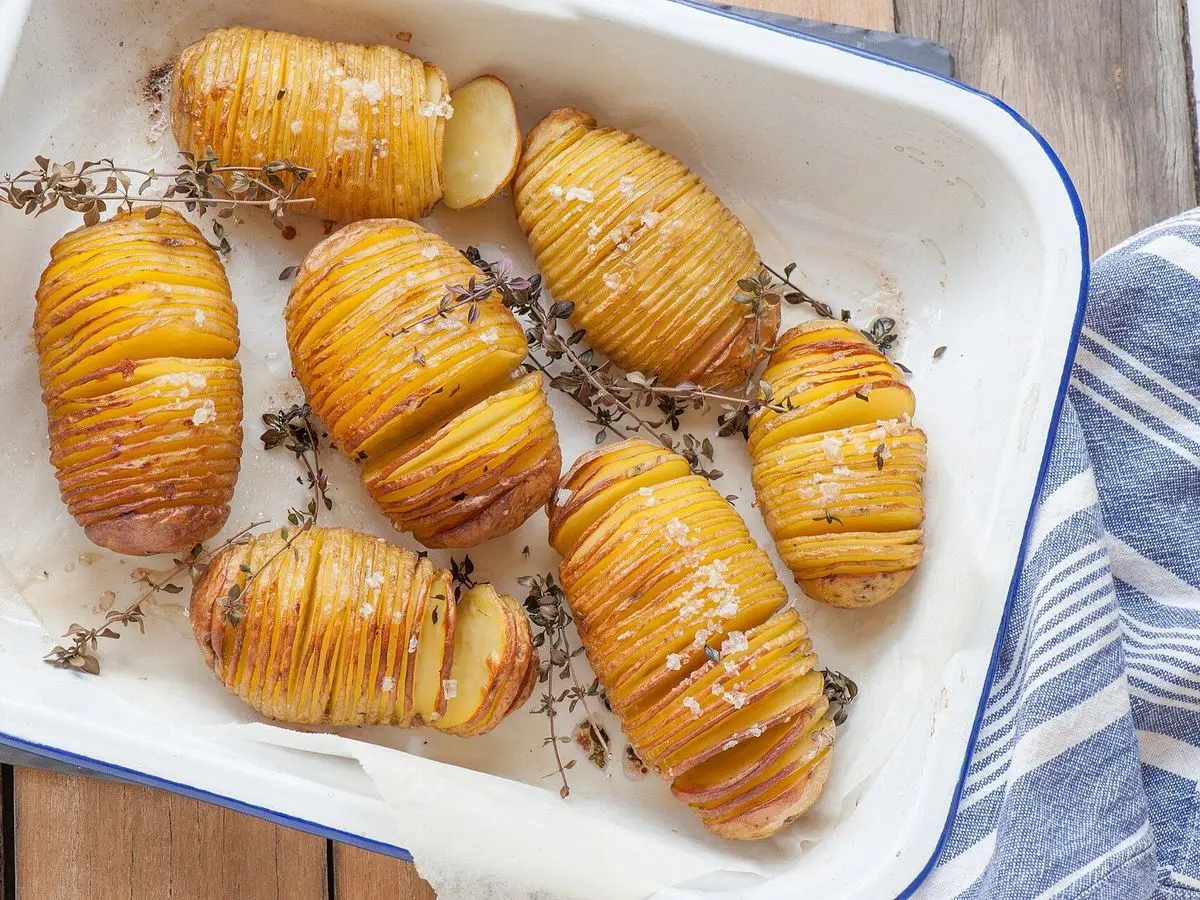 Patatas hasselback laminadas y doradas con tomillo en una fuente esmaltada blanca