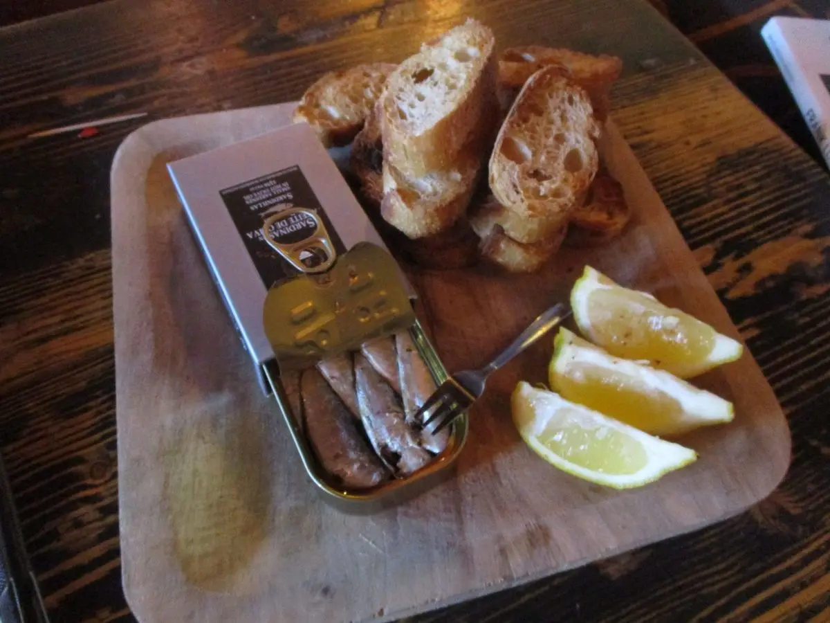 Lata abierta de sardinas junto a trozos de pan y gajos de limón sobre una tabla de madera