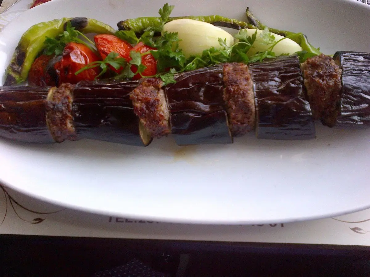 Brocheta de berenjena asada intercalada con carne picada, con tomate, pimiento y yogur en un plato blanco