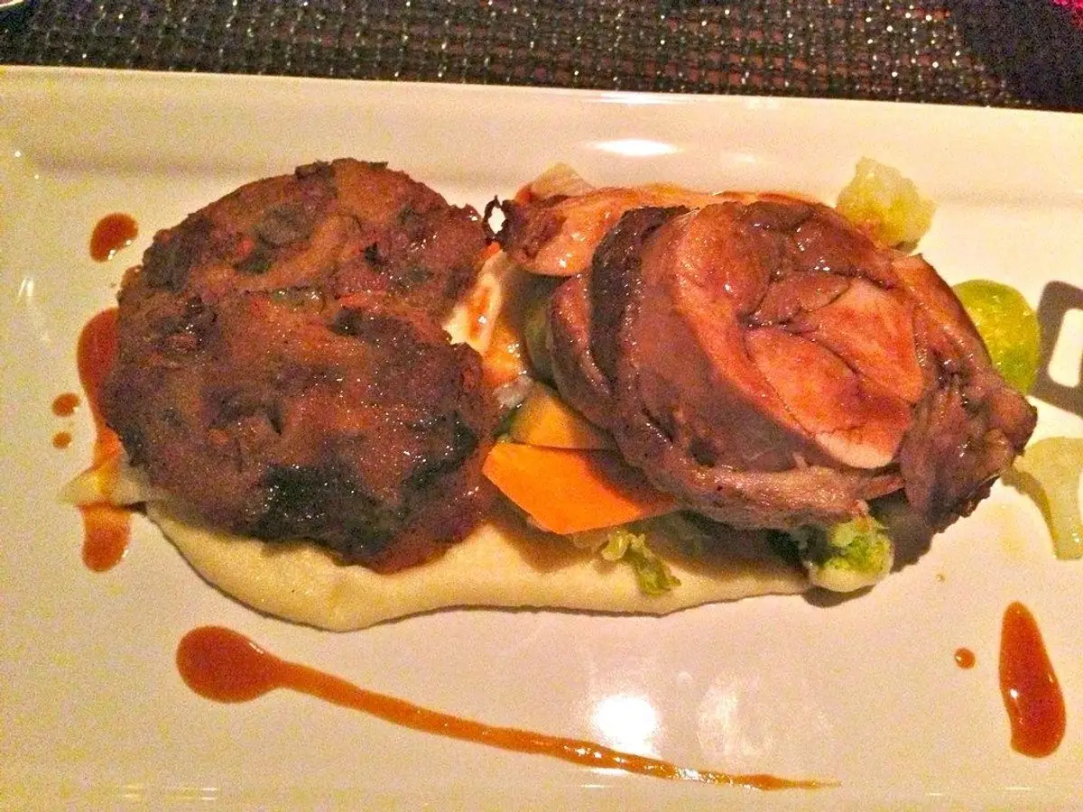 Dos piezas de carne doradas, una enrollada, sobre puré cremoso con verduras y salsa roja en un plato blanco
