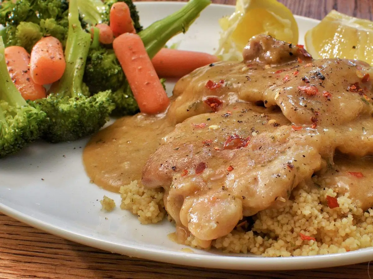 Pechuga de pollo en salsa sobre cuscús con brócoli y zanahoria en un plato blanco