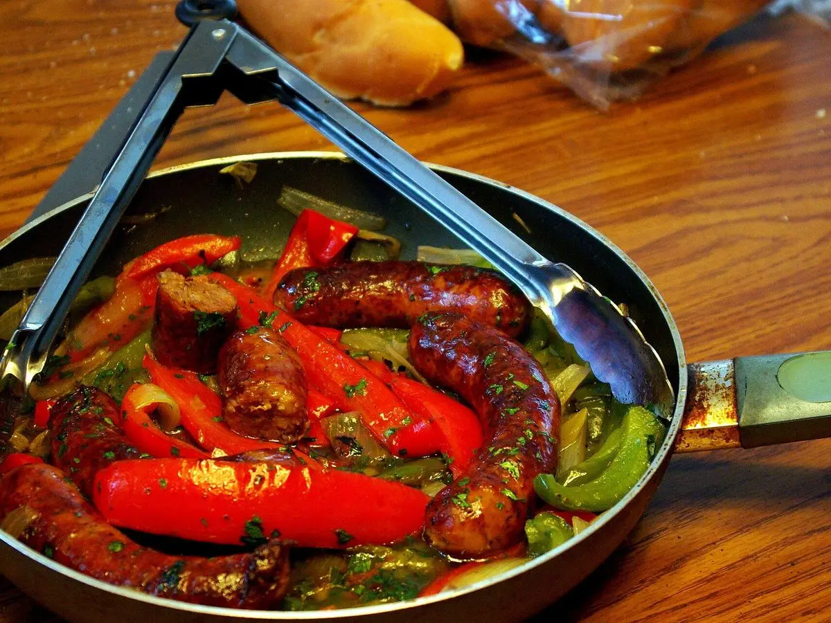 Sartén con salchichas asadas y tiras de pimiento rojo y verde, con unas pinzas
