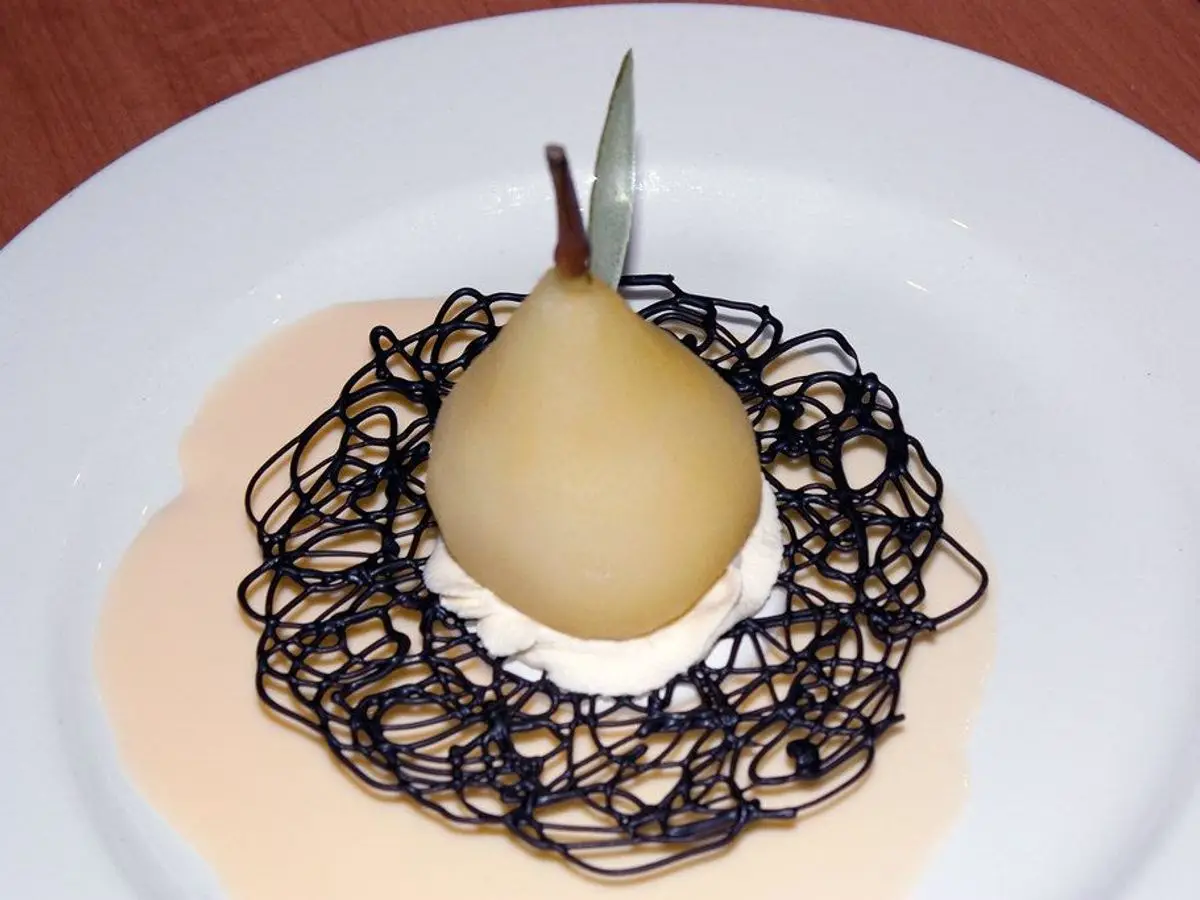 Pera pochada de tono claro sobre un plato blanco con crema y un adorno de chocolate