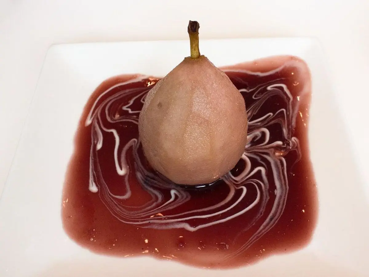Pera entera escalfada en vino o té rojo sobre un plato blanco con salsa granate en remolinos