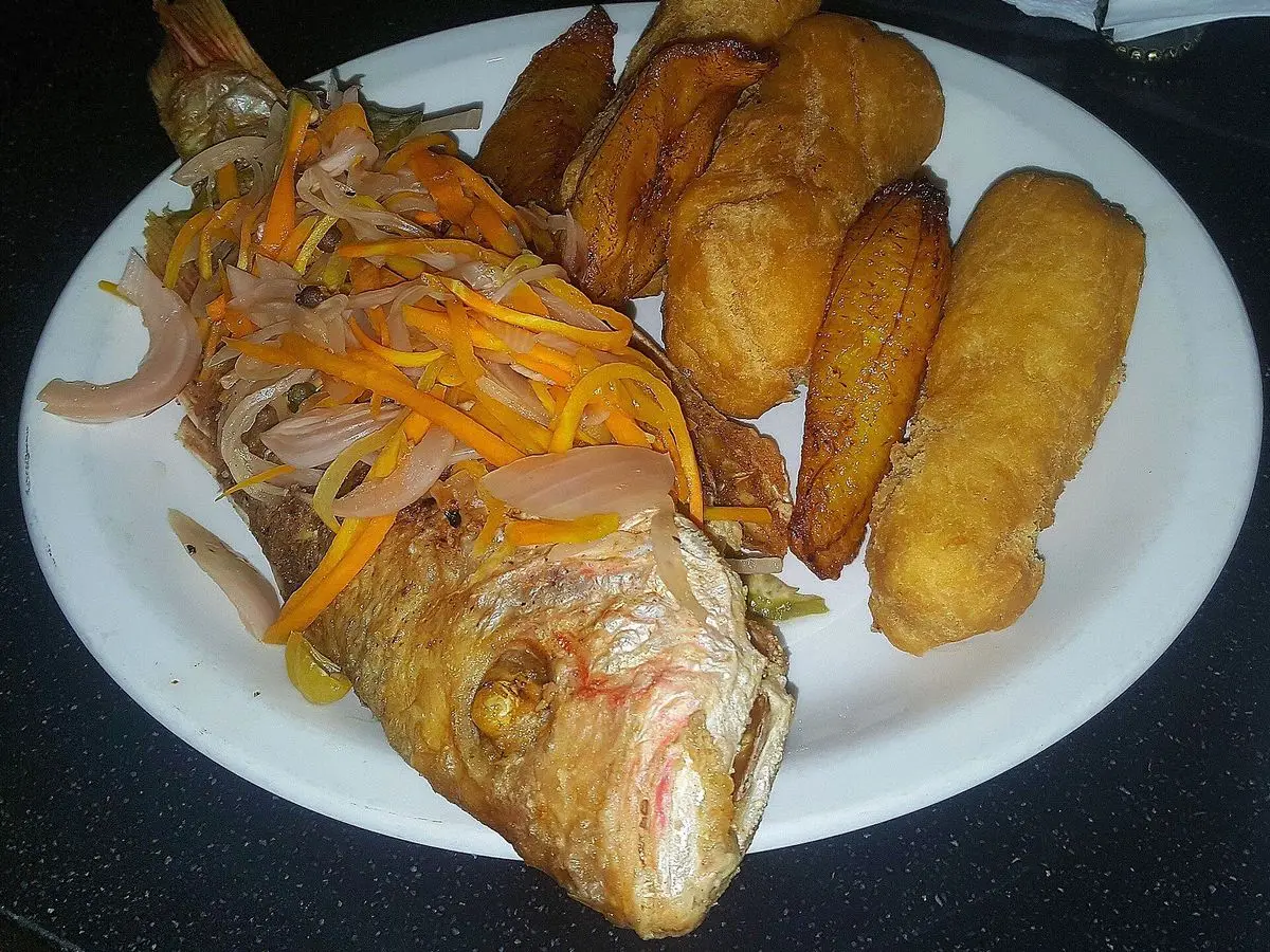 Pescado entero frito con cebolla y zanahoria en tiras, acompañado de fritos de masa dorados en un plato blanco