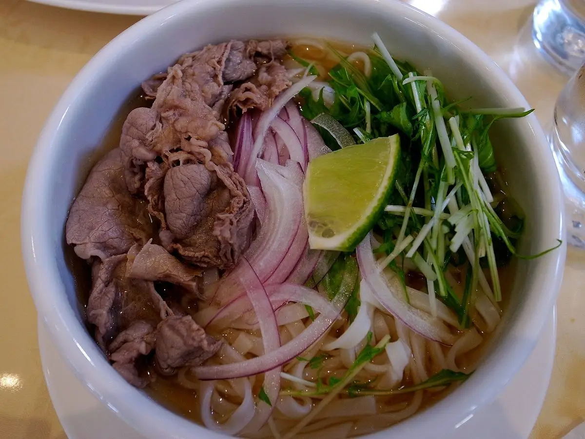 Cuenco de sopa pho con lonchas de ternera, cebolla morada, brotes, hierbas y una rodaja de lima