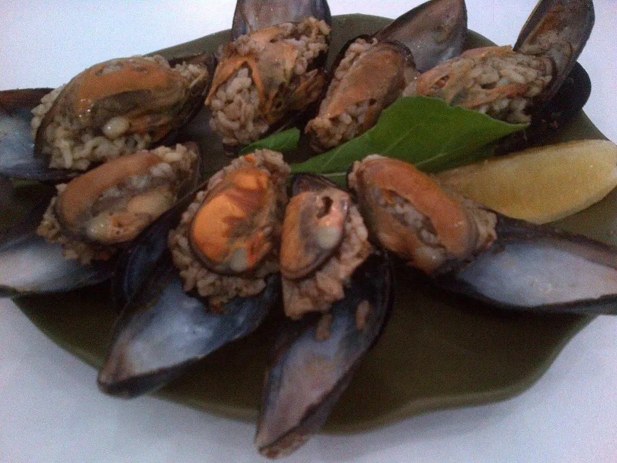 Plato verde con mejillones rellenos de arroz especiado dispuestos en círculo con una cuña de limón