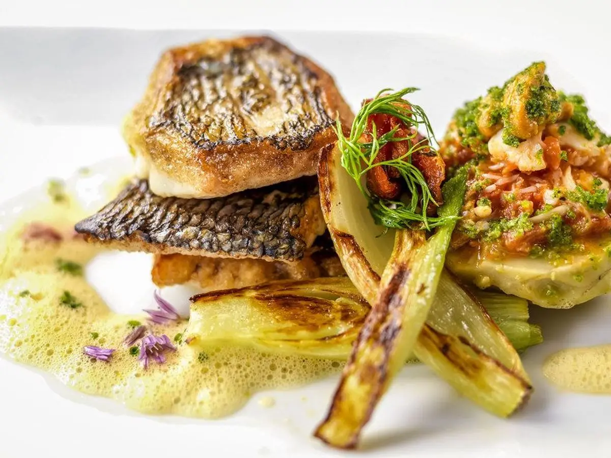 Lubina con piel crujiente sobre verduras salteadas y salsa amarilla en un plato blanco