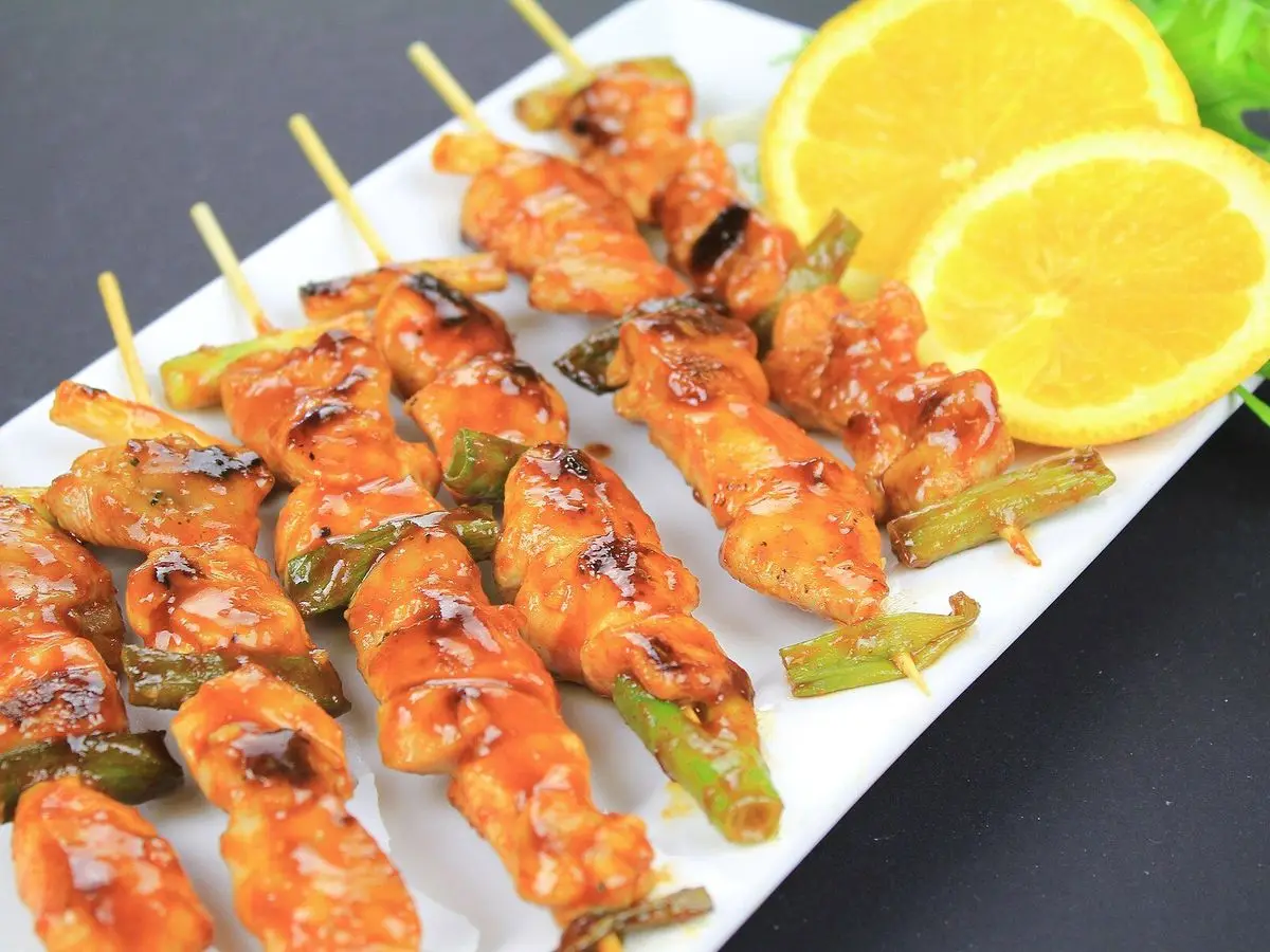 Pinchos de pollo especiados dorados con trozos de cebolleta sobre una fuente blanca con rodajas de naranja