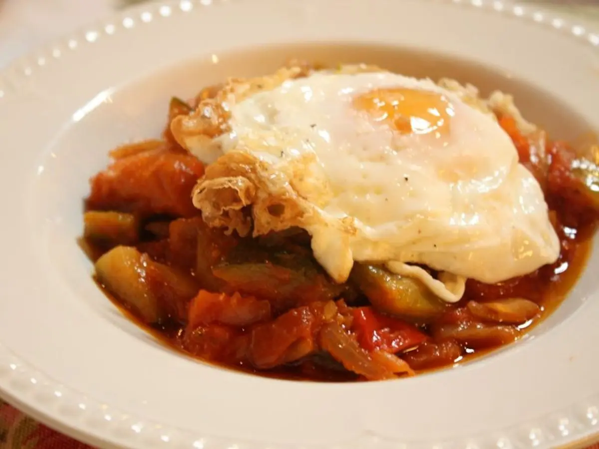 Pisto de tomate, pimiento y calabacín con un huevo frito encima en un plato hondo blanco