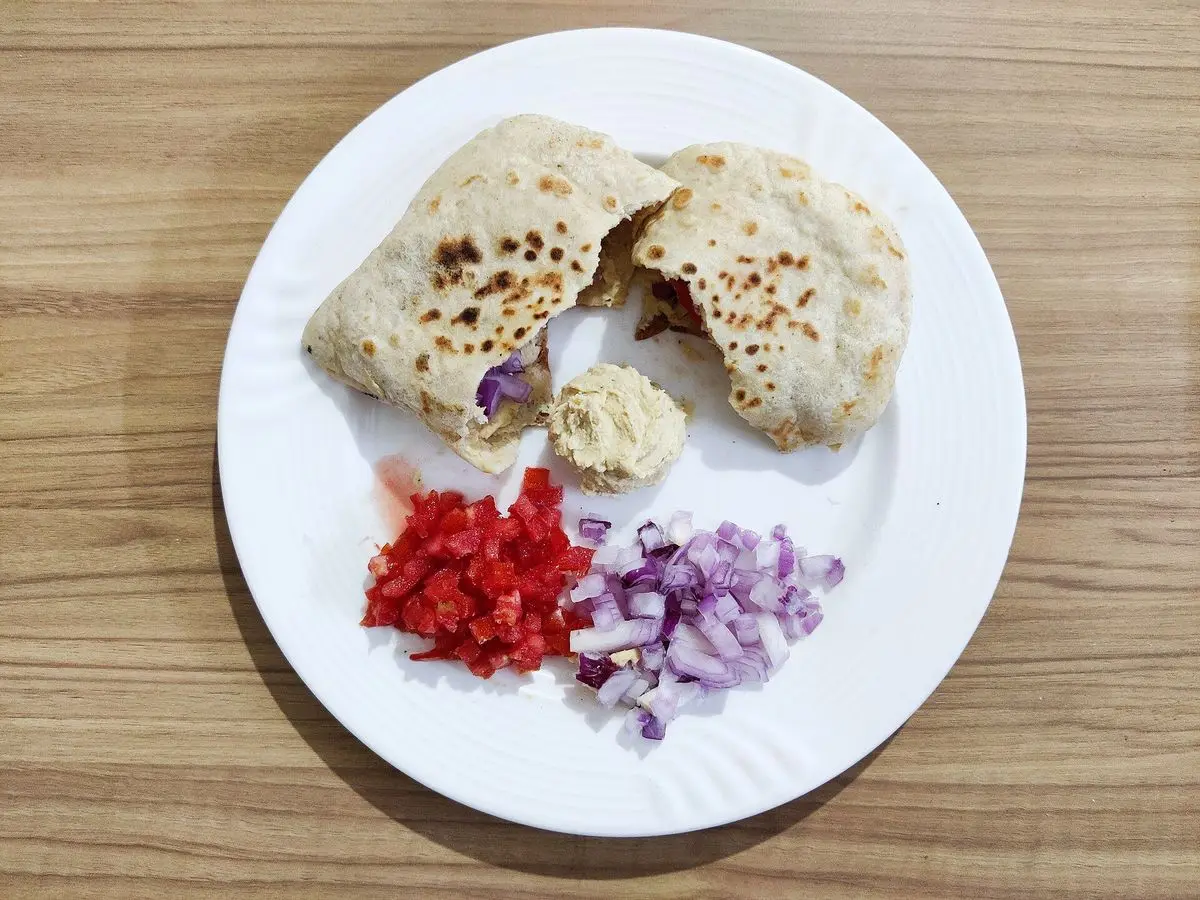 Pan de pita relleno con hummus junto a tomate picado y cebolla morada en un plato blanco