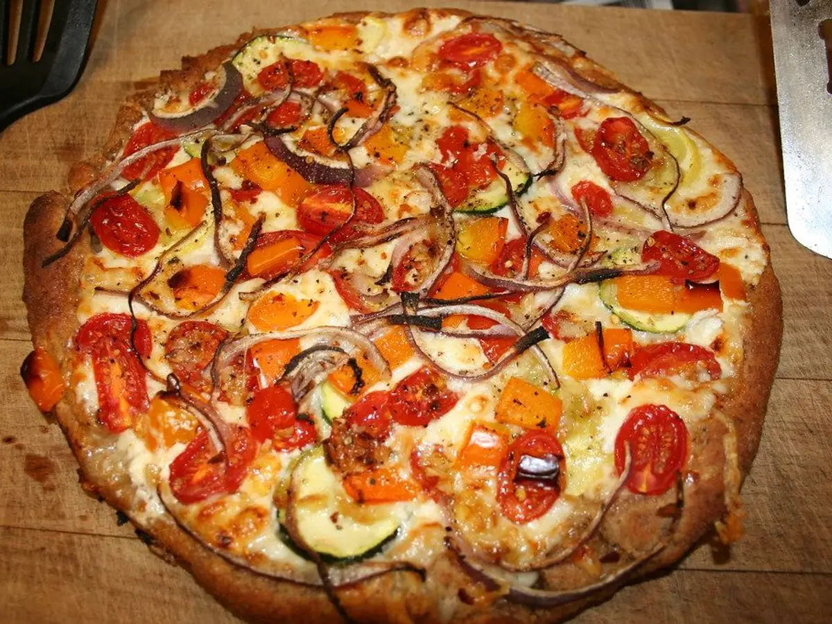 Pizza casera de masa fina con rodajas de calabacín, pimiento, cebolla roja, tomate y queso sobre tabla de madera