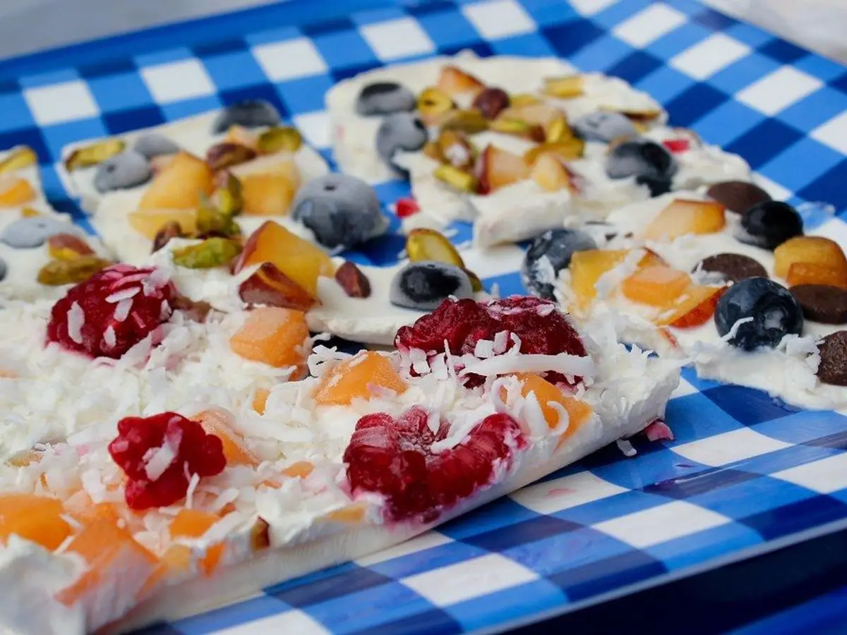 Placa helada de yogur con frutos rojos, mango, coco rallado y frutos secos sobre un mantel de cuadros azules