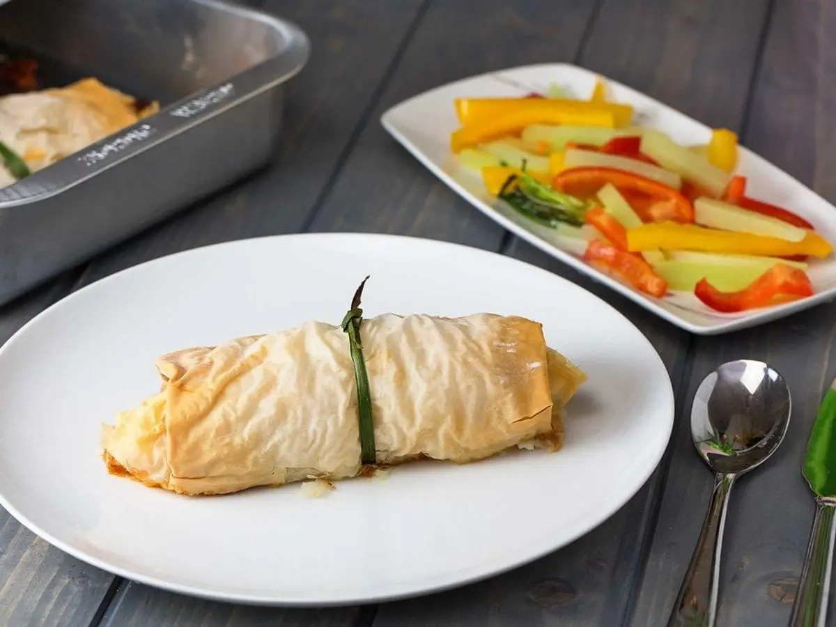Pechuga de pollo rellena enrollada y dorada en un plato blanco, con una guarnición de verduras al fondo