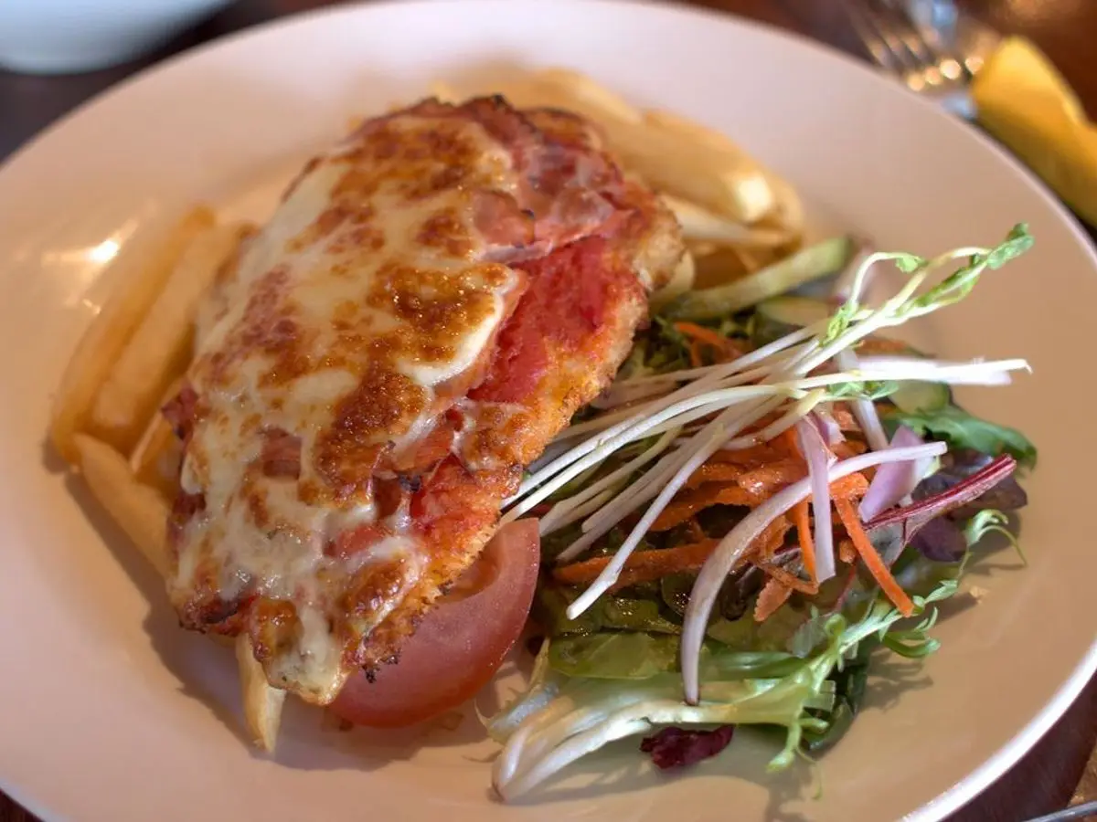 Filete de pollo empanado gratinado con queso y tomate junto a ensalada y patatas fritas en un plato blanco