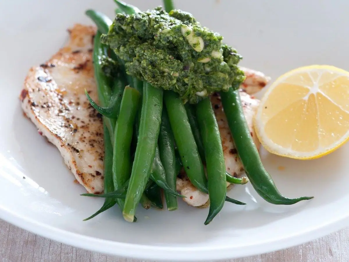 Pechuga de pollo a la plancha con judías verdes cubiertas de pesto y medio limón en un plato blanco