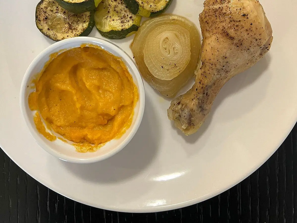 Plato blanco con muslo de pollo asado, calabacín a la plancha, cebolla y cuenco de salsa anaranjada