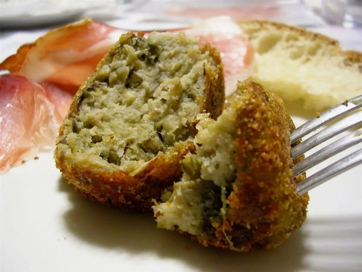 Albóndiga vegetal rebozada partida por la mitad con relleno verde, con un tenedor y pan al fondo