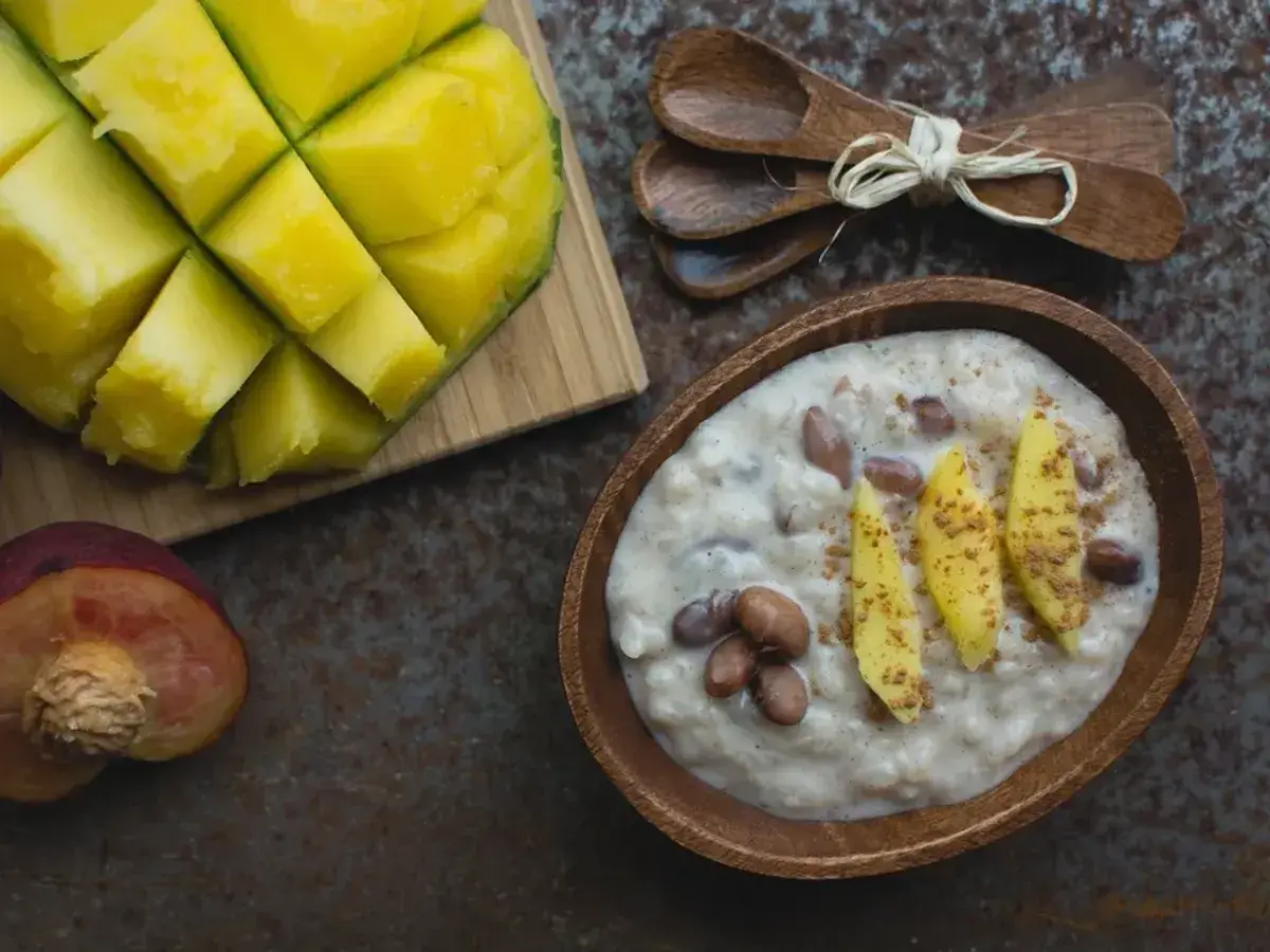 Cuenco de madera con gachas cremosas de coco, láminas de mango y canela, junto a un mango cortado en dados