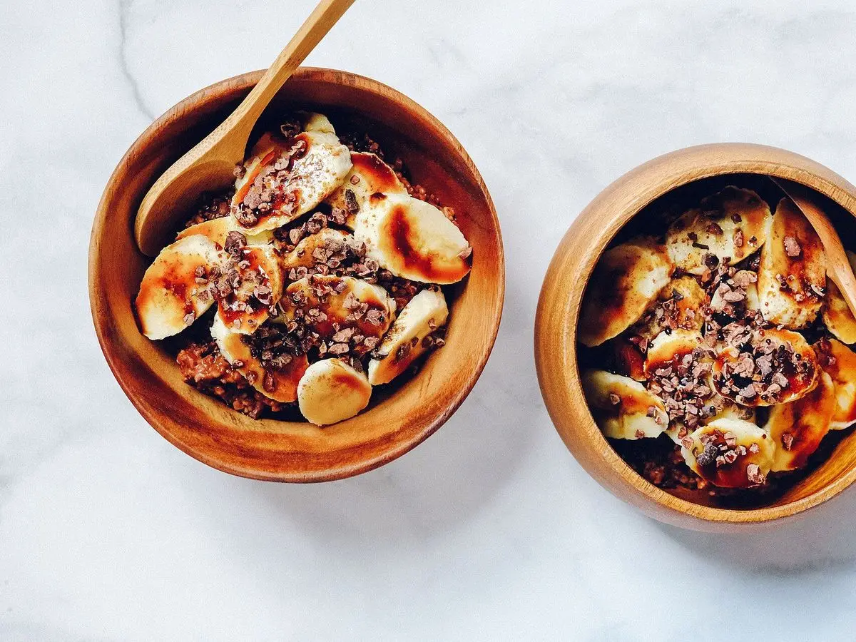 Dos cuencos de madera con avena, rodajas de plátano y cacao sobre una mesa blanca