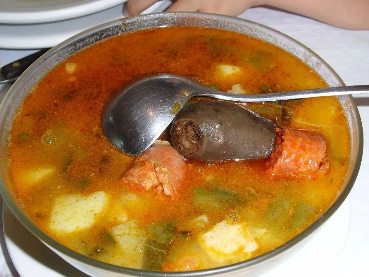Fuente de pote con berza, patata, chorizo y morcilla en caldo anaranjado con un cucharón