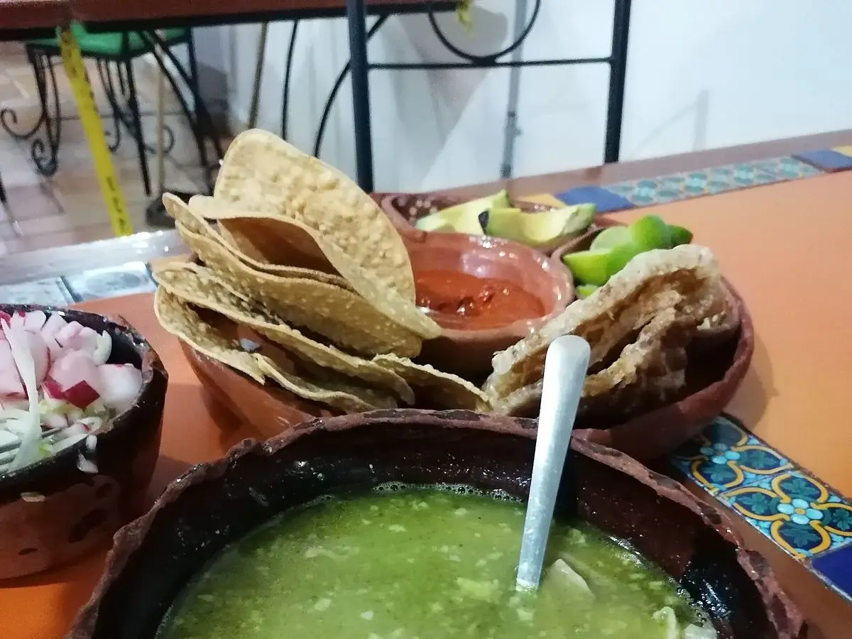 Cuenco de barro con pozole verde y una cuchara, con tostadas y salsa al fondo