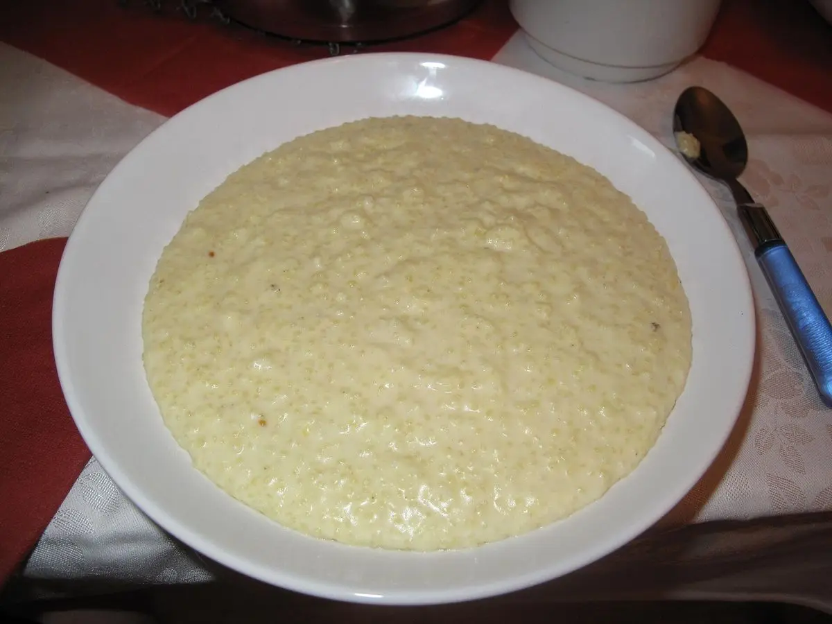 Plato hondo blanco con gachas cremosas de mijo cocido, junto a una cuchara sobre un mantel