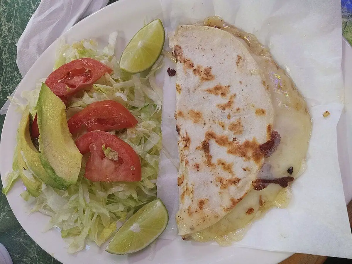 Quesadilla de queso fundido doblada junto a lechuga, tomate, aguacate y lima en un plato blanco