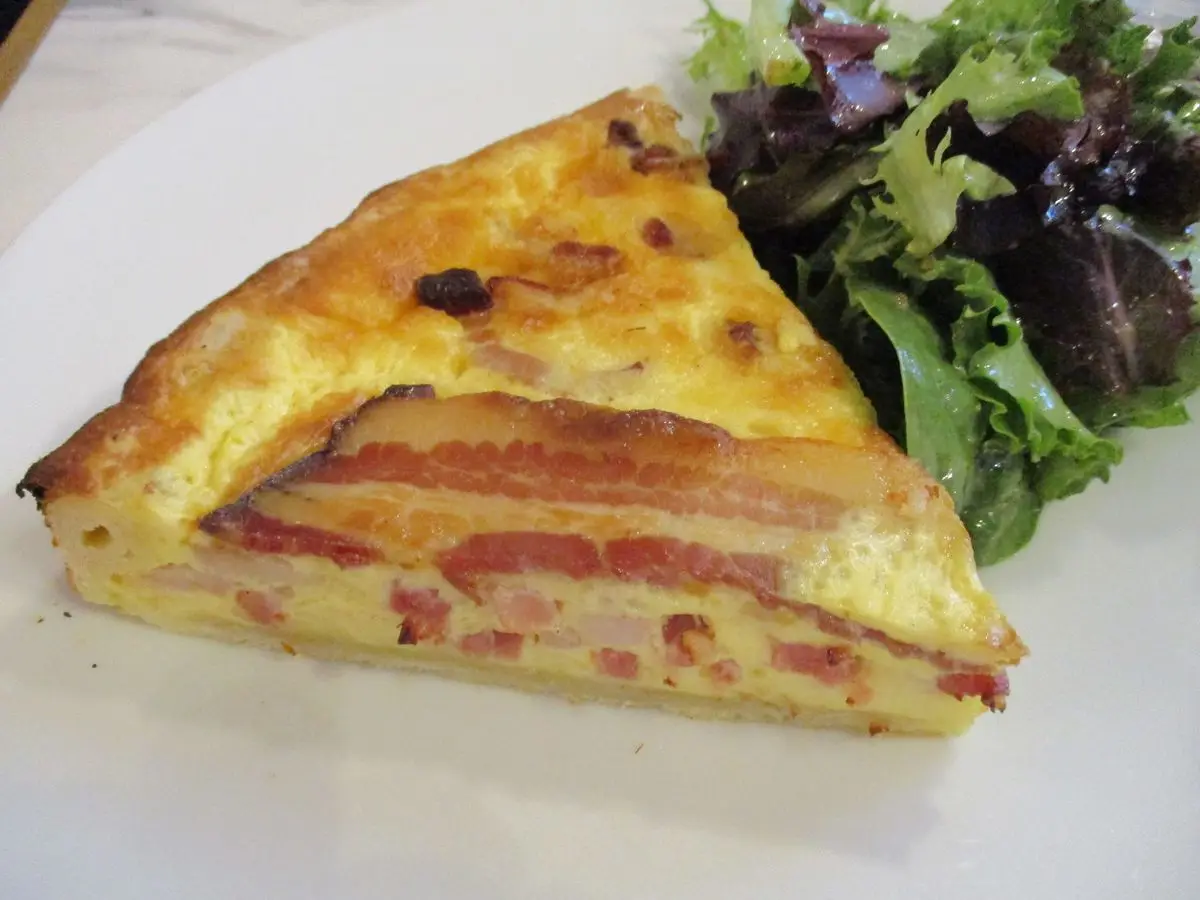 Porción de quiche con jamón y tomate junto a ensalada de hojas verdes en un plato blanco