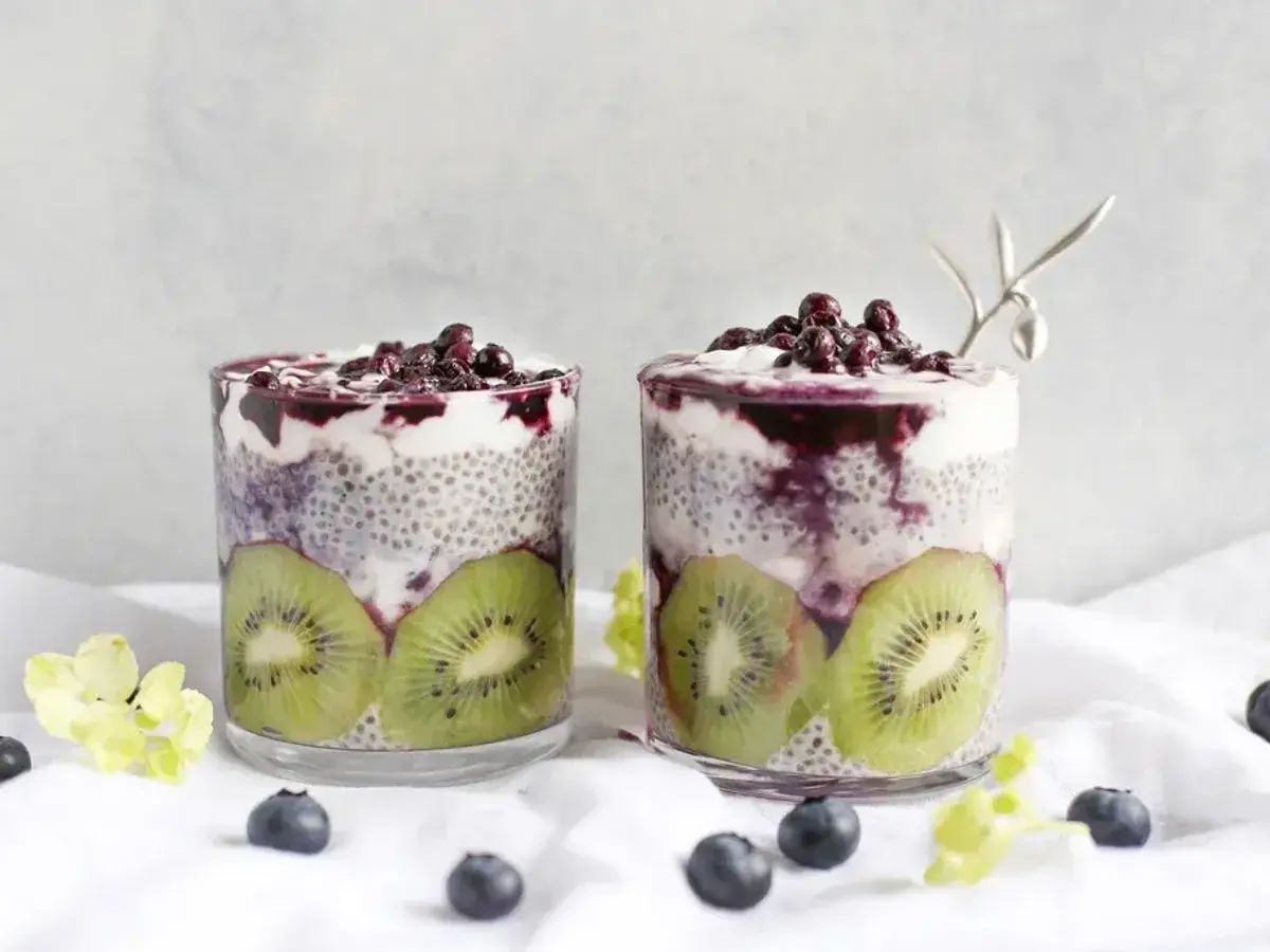 Dos vasos con capas de pudin de chía, yogur y rodajas de kiwi, coronados con arándanos