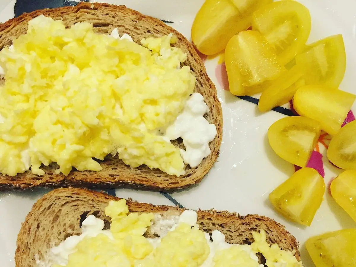 Rebanadas de pan integral con huevo revuelto y un poco de queso blanco, junto a tomates amarillos troceados