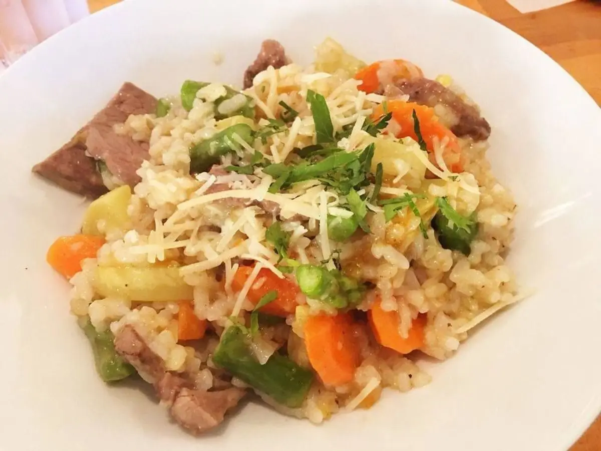Plato blanco de risotto con trozos de cordero, zanahoria, verduras y queso rallado