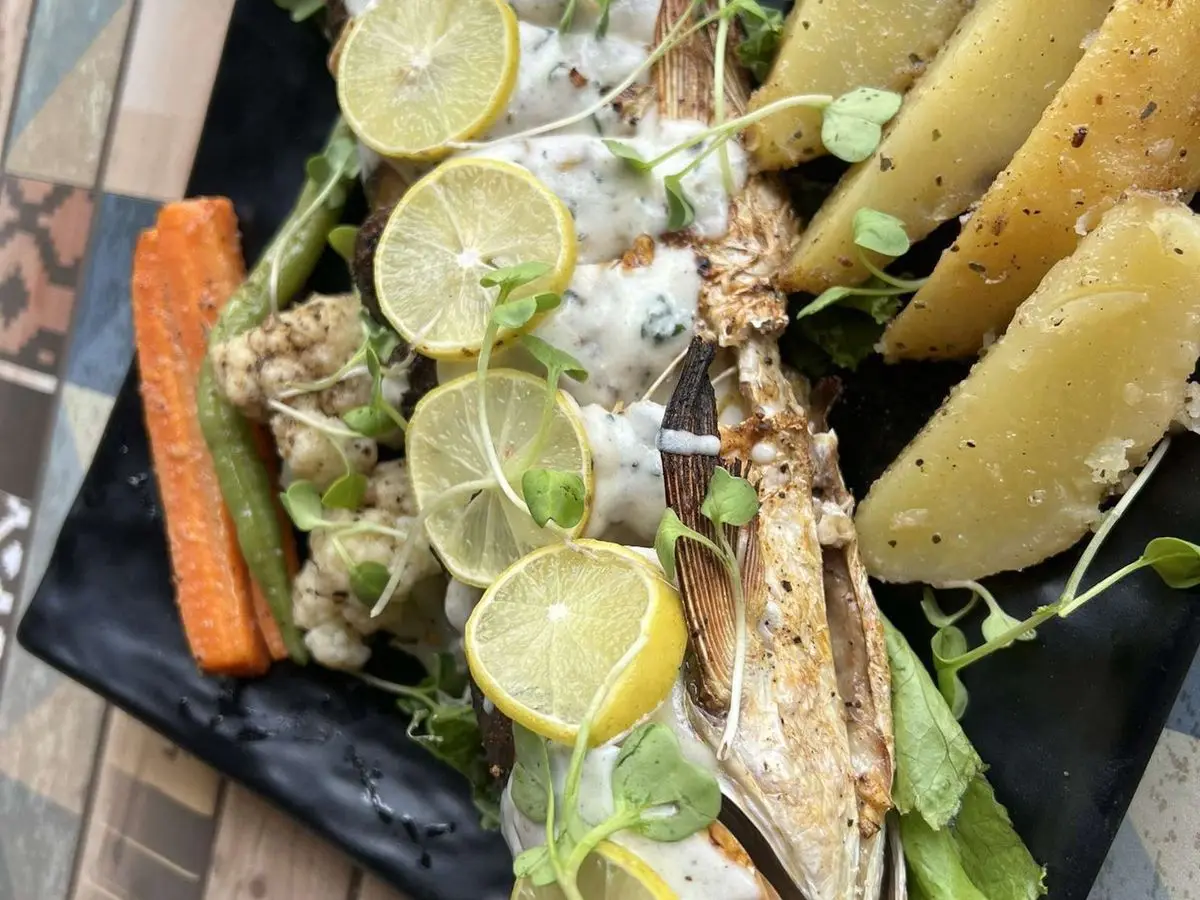 Pescado entero a la brasa con rodajas de limón, patatas asadas, lechuga y zanahoria sobre una fuente negra
