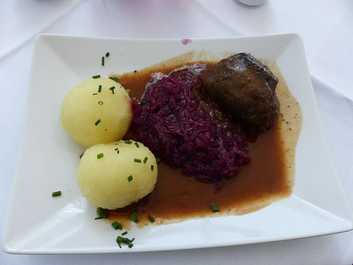 Rollo de ternera con salsa, lombarda y dos patatas cocidas con cebollino en un plato cuadrado