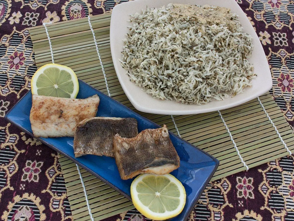 Arroz persa con hierbas verdes en un plato blanco junto a filetes de pescado dorado y rodajas de limón
