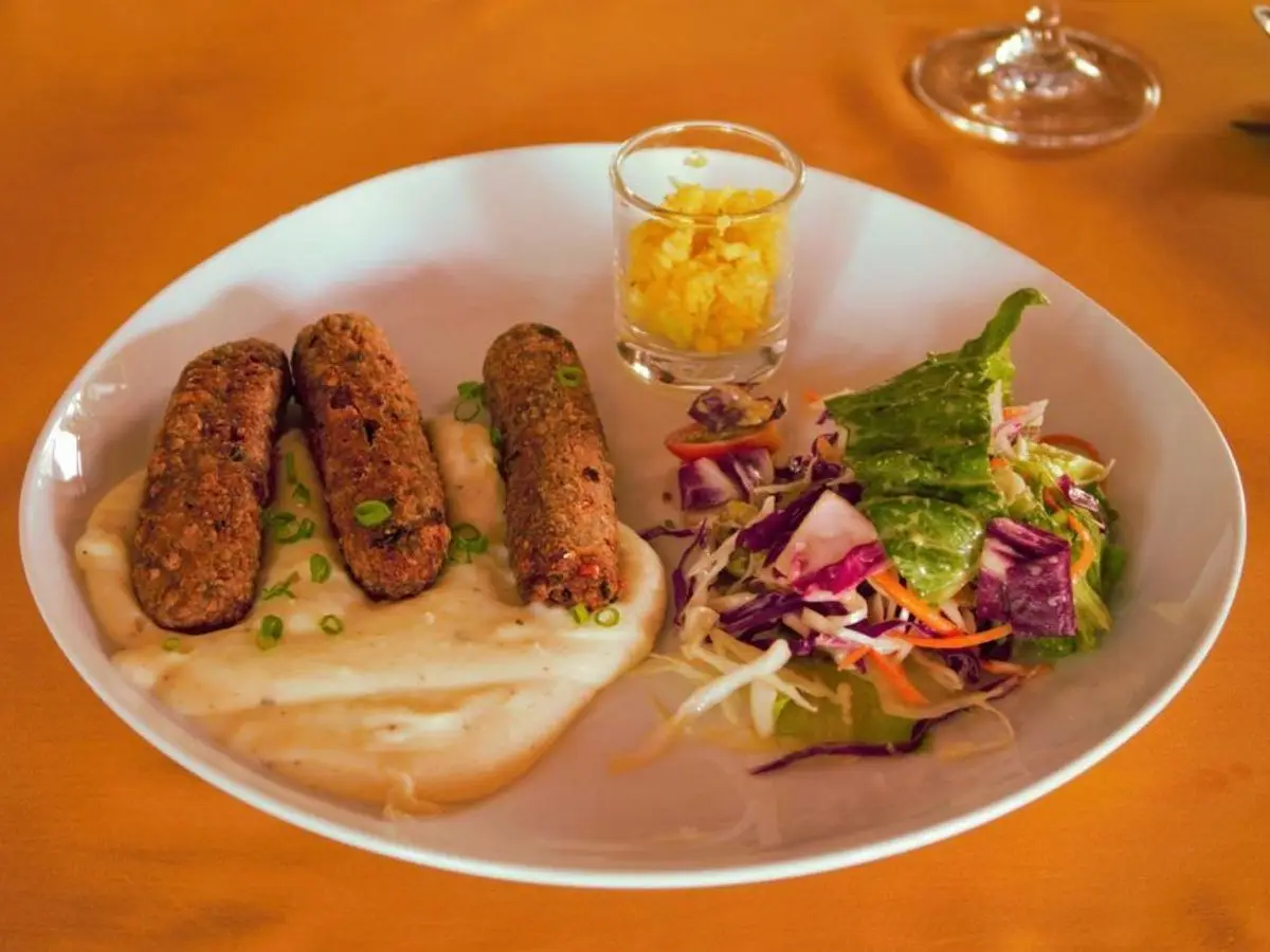 Tres salchichas veganas doradas sobre puré de patata cremoso con ensalada de col morada en un plato blanco