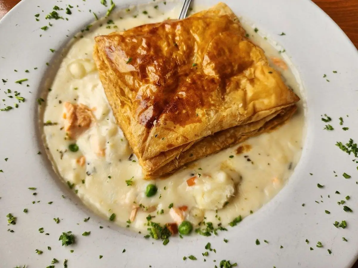 Porción de hojaldre dorado sobre salsa cremosa con guisantes y hierbas en un plato blanco