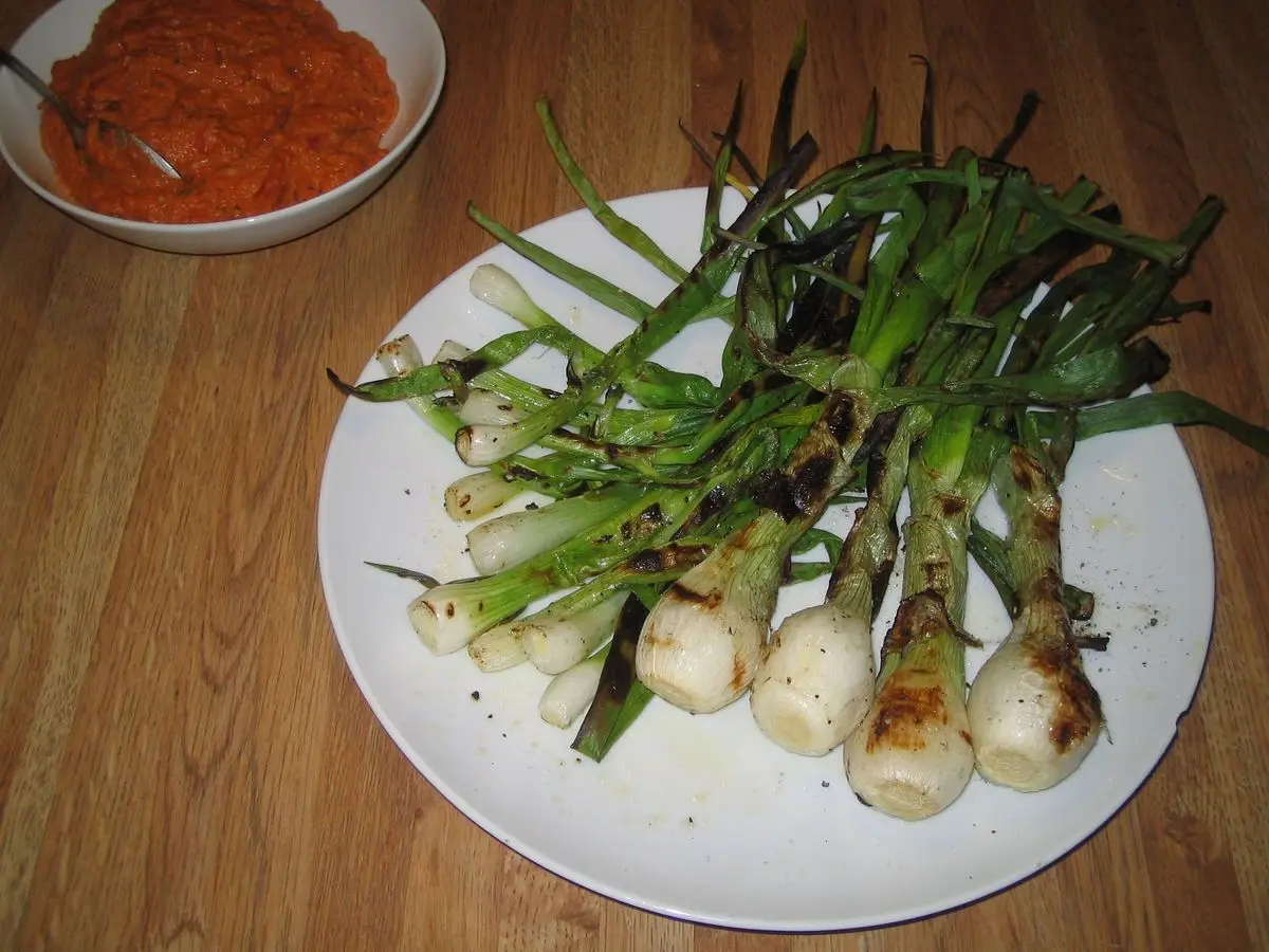Plato con calçots a la brasa y un cuenco de salsa romesco naranja al fondo sobre una mesa de madera