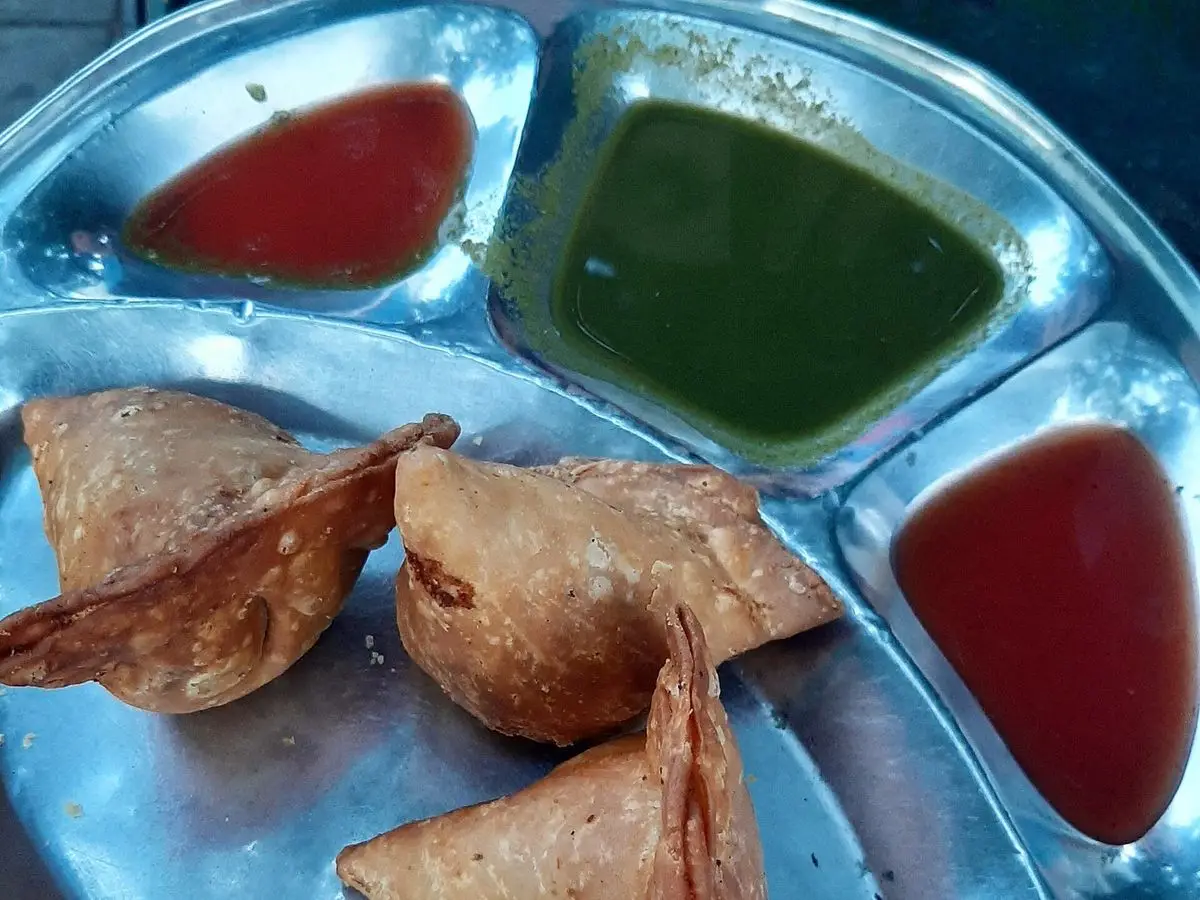 Samosas triangulares doradas en una bandeja metálica con chutney verde y salsa roja