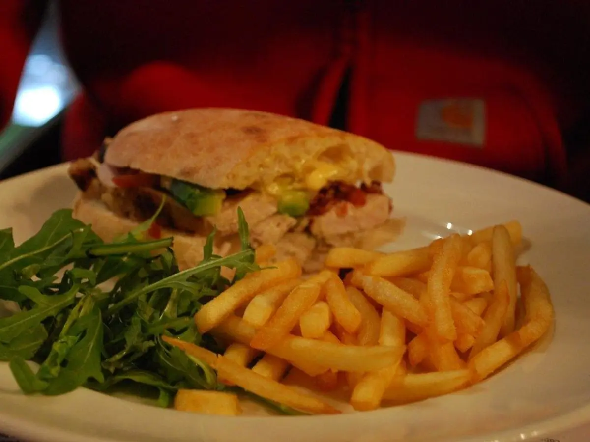 Sándwich en pan redondo con pollo y hojas verdes, acompañado de patatas fritas y rúcula