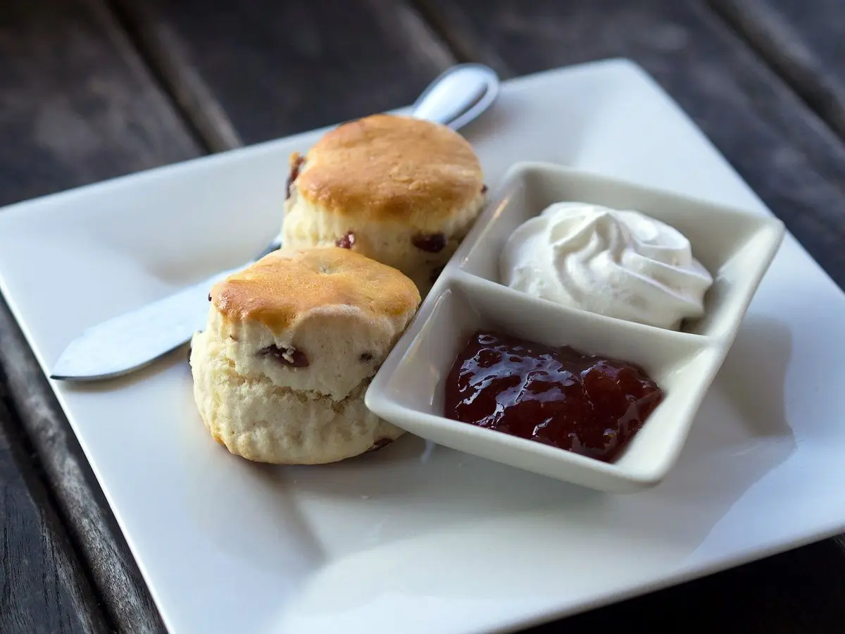 Dos scones con un cuenco de nata montada y otro de mermelada roja en un plato blanco