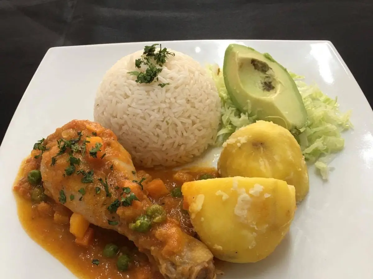 Muslo de pollo guisado en salsa de tomate con arroz blanco, aguacate y patata en un plato blanco