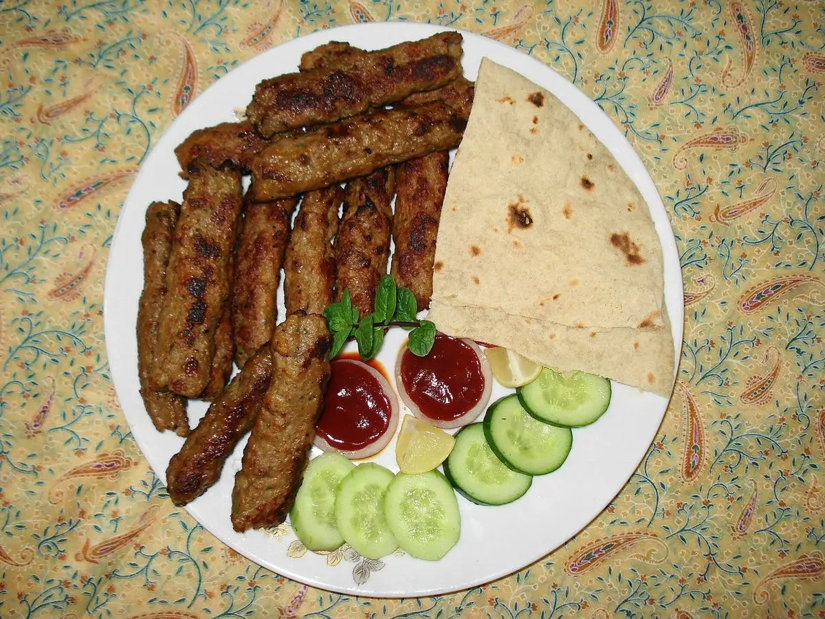Plato de seekh kebab asado con pan plano, rodajas de pepino y salsas rojas sobre un mantel estampado