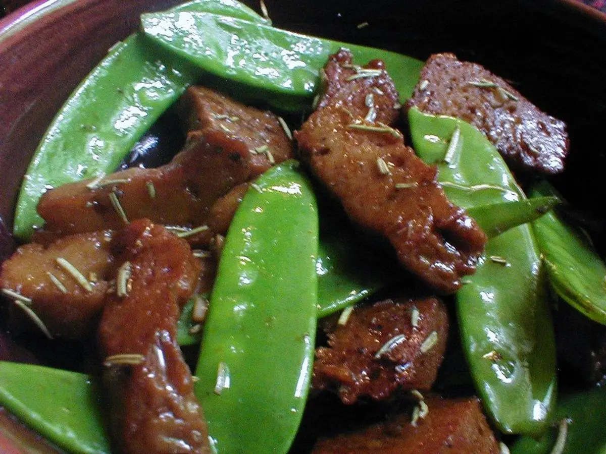 Trozos de seitán dorado salteados con tirabeques verdes y romero en un plato oscuro