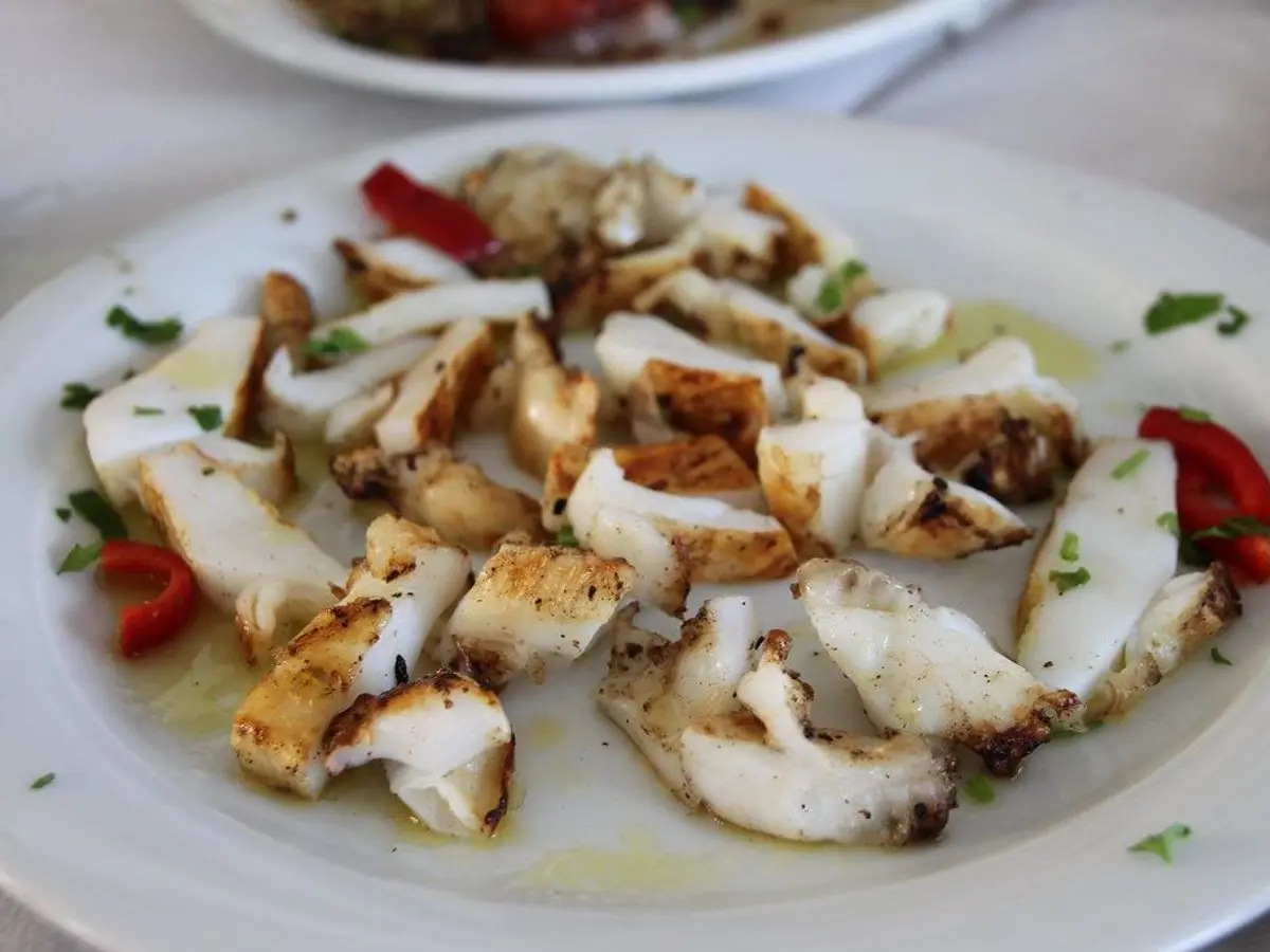 Plato blanco con trozos de sepia o calamar a la plancha, pimiento rojo y hierbas, con un hilo de aceite