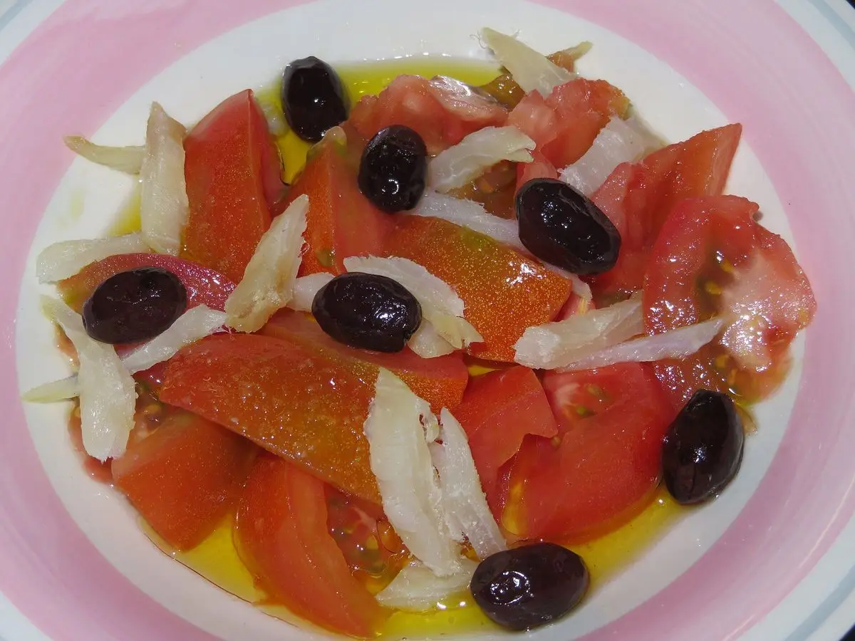 Ensalada de tomate con migas de bacalao desmenuzado, cebolla y aceitunas negras en un plato rosa y blanco