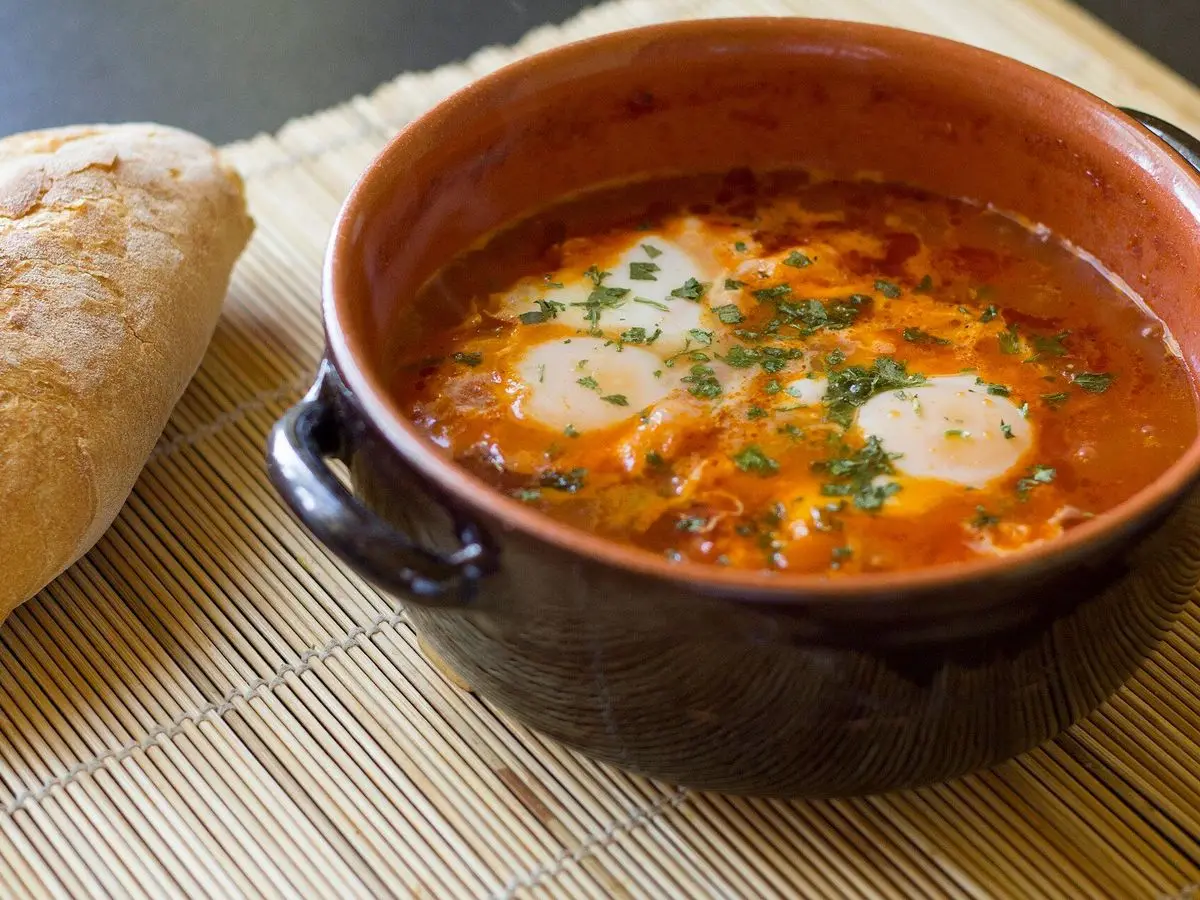 Cuenco de barro con shakshuka, huevos en salsa de tomate con hierbas, junto a una hogaza de pan