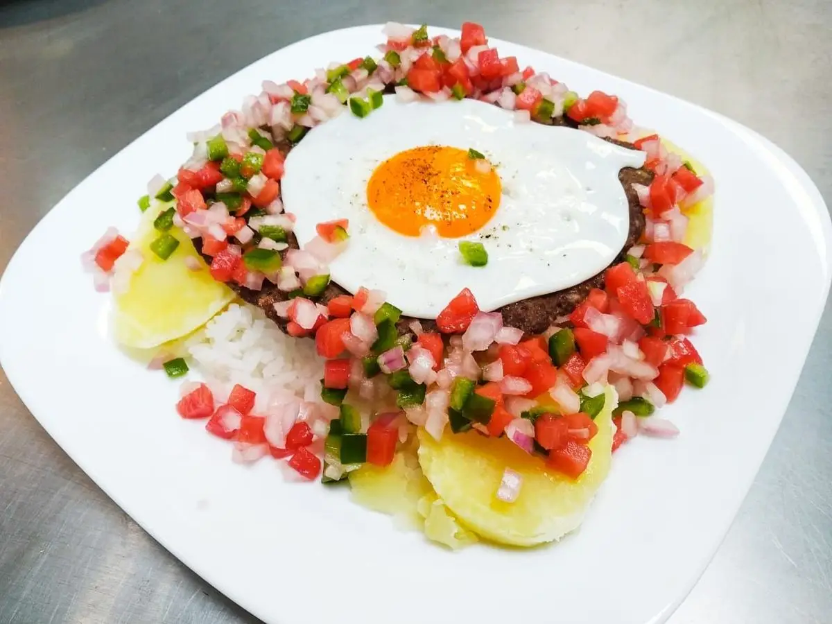 Silpancho con huevo frito, salsa cruda de tomate y cebolla, patata y arroz en un plato blanco